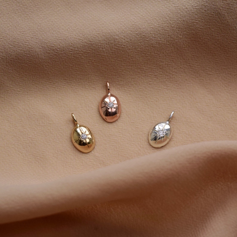 May include: Three small, oval pendants in gold, rose gold, and silver. Each pendant has a starburst design with a small, clear gemstone in the centre. The pendants are displayed on a neutral, tan-coloured fabric.