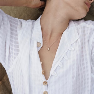 May include: A delicate gold necklace with a small, oval pendant. The pendant hangs on a thin chain and is centered on a person's chest. The person is wearing a white, button-down shirt with a subtle striped pattern.