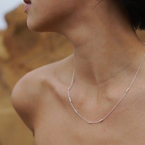 May include: A silver necklace with small, evenly spaced beads. The necklace is worn by a person with a bare neck and shoulders.