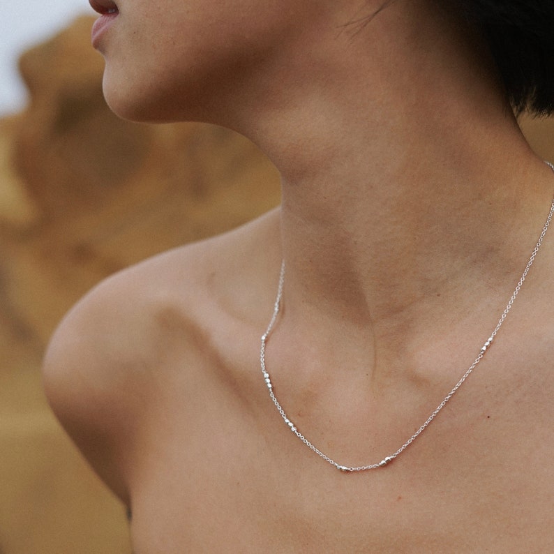 May include: A silver necklace with small, evenly spaced beads. The necklace is worn by a person with a bare neck and shoulders.
