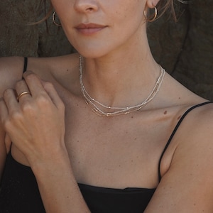 May include: A woman wearing a black top with a layered gold and silver necklace. The necklace is made of small beads and is worn close to the neck.
