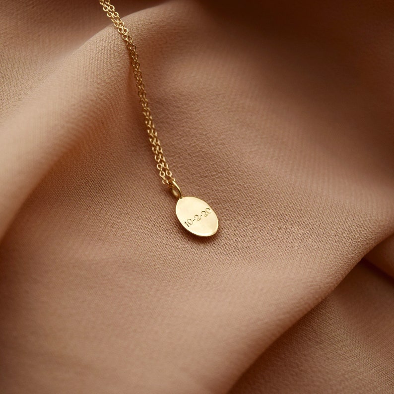 May include: A gold necklace featuring a fine chain and an oval pendant. The pendant is engraved with the date "10-2-20". The necklace is presented against a backdrop of soft, tan fabric, offering a minimalist and refined appearance.