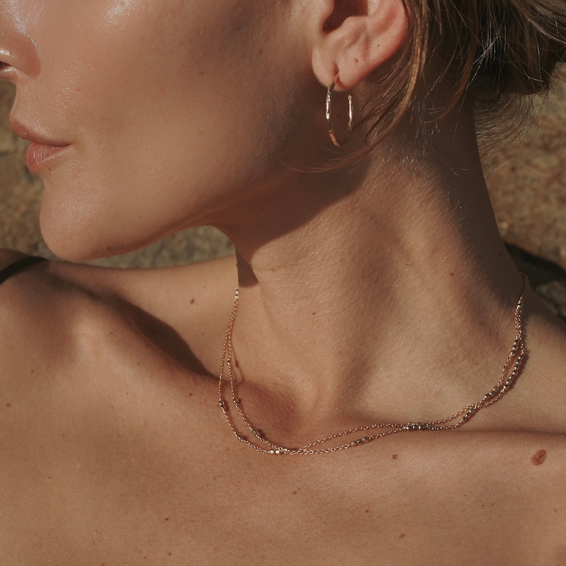 May include: A close-up of a person wearing a gold necklace with two delicate chains. The necklace features small, round beads. The person is also wearing a gold hoop earring.