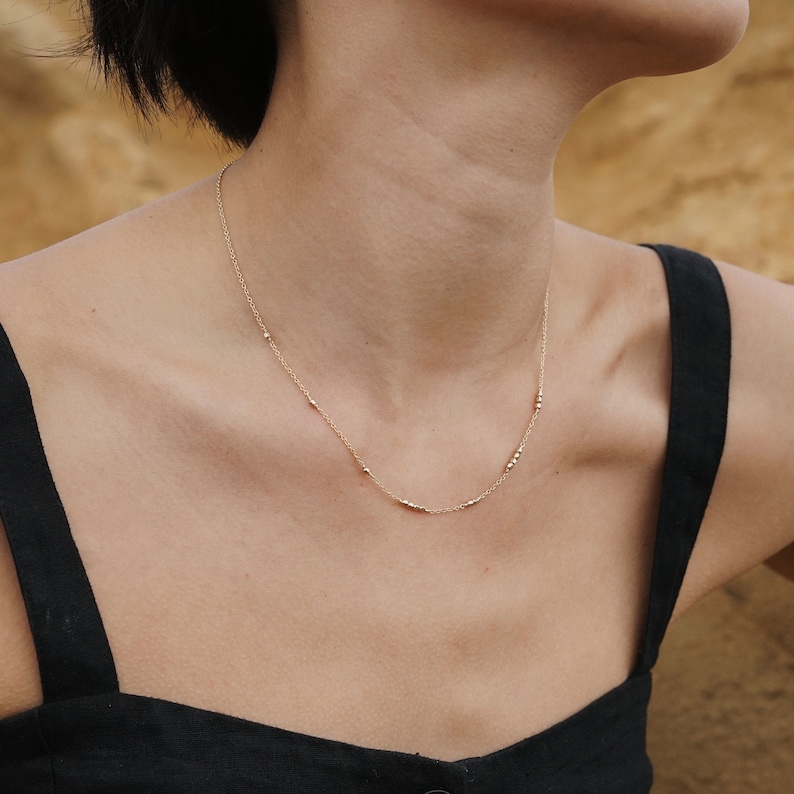 May include: A delicate gold necklace with small beads, worn by a person with short dark hair.