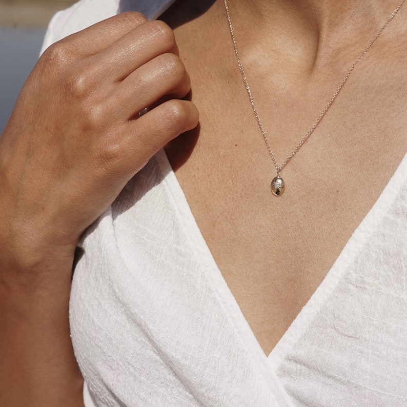 May include: A gold pendant necklace featuring a small, oval charm with a textured surface. The charm hangs from a fine chain. The necklace is worn against a white top.