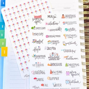May include: A planner with stickers featuring colorful balloons and holiday-themed text. The stickers include phrases like "Happy New Year," "Valentine's," and "Christmas." The planner has gold spiral binding and colorful tabs.