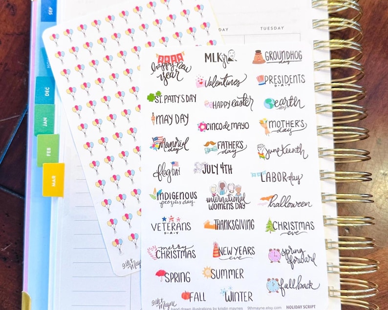 May include: A planner with stickers featuring colorful balloons and holiday-themed text. The stickers include phrases like "Happy New Year," "Valentine's," and "Christmas." The planner has gold spiral binding and colorful tabs.