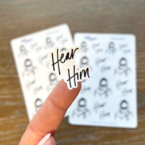 Peut inclure: Une feuille d'autocollants avec un fond blanc et des contours noirs de personnes. Les autocollants ont les mots "Hear Him" &eacute;crits en noir.