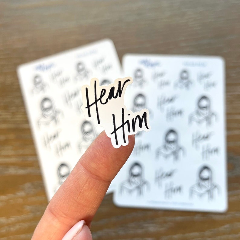 Peut inclure: Une feuille d'autocollants avec un fond blanc et des contours noirs de personnes. Les autocollants ont les mots "Hear Him" &eacute;crits en noir.