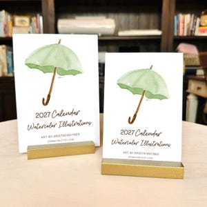 May include: Two white rectangular cards featuring a watercolor illustration of a light green umbrella with a brown handle. The text reads 2027 Calendar Watercolor Illustrations, Art by Kristin Maynes, 8thmayne.etsy.com. Each card sits in a gold stand.
