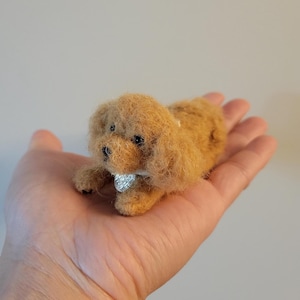 May include: A small, handmade, tan-colored dog figurine with a fluffy texture. The dog is lying down and wearing a silver collar. The figurine is resting in the palm of a hand.
