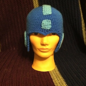 May include: A blue crocheted hat with a helmet design and blue accents. The hat is on a mannequin head.