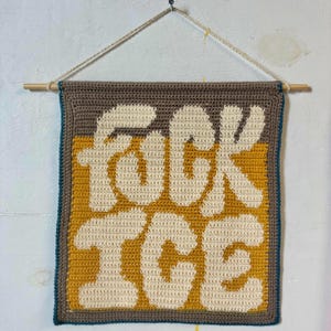 Peut inclure: Une tenture murale au crochet faite à la main avec les mots "FUCK ICE" en grandes lettres blanc cassé. Le texte est placé sur un fond jaune moutarde, encadré d'une bordure marron et d'un bord turquoise. Une cheville en bois et une ficelle sont utilisées pour l'accrocher.