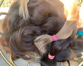 以下が含まれることがあります： 茶色とブロンドの様々な色合いのヘアエクステンションのコレクション。髪は束ねられ、赤、青、ピンクなどのカラフルな輪ゴムで固定されています。画像は、自然な質感と色のバリエーションを示しています。