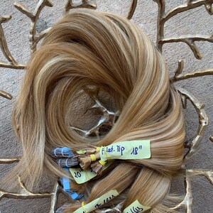 May include: A bundle of blonde and light brown hair extensions with highlights. The extensions are labelled with lengths of 45.7 and 50.8 cm. The hair is arranged in a circular pattern.
