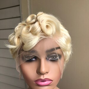 Puede incluir: Una peluca rubia corta con una parte delantera de encaje. La peluca presenta secciones de cabello muy rizado. La peluca está peinada en una cabeza de maniquí con un tono de piel neutro. La peluca es de color rubio dorado claro.