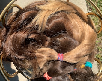 以下が含まれることがあります： 茶色とブロンドの様々な色合いのヘアエクステンションのコレクションで、カラフルな輪ゴムで固定されています。髪は円形に配置され、さまざまな色と質感を表現しています。画像は自然光で撮影されています。