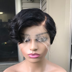 Può includere: Una parrucca nera con taglio pixie e frontale in pizzo è esposta su una testa di manichino. La parrucca presenta capelli corti e mossi acconciati di lato. Il manichino ha labbra rosa e un incarnato neutro.