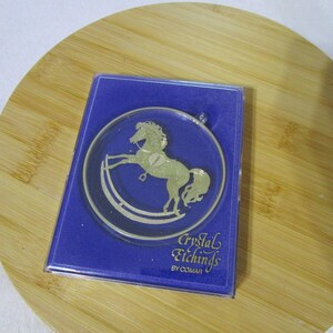 May include: A blue box with a clear round ornament featuring a gold rocking horse. The ornament is labeled "Crystal Etchings by Comar".