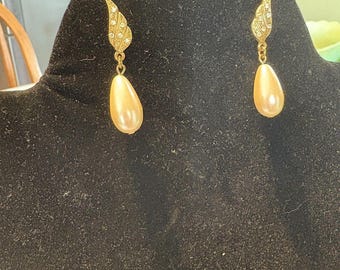 May include: A pair of gold-toned drop earrings. Each earring features a decorative, wing-shaped top encrusted with small, clear crystals. A pear-shaped, cream-colored faux pearl dangles below, creating an elegant, classic look.