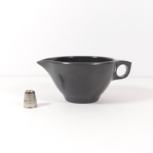 May include: A dark gray ceramic creamer with a handle. The creamer is sitting on a white surface.