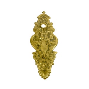 May include: A gold-toned metal door plate with an ornate design. The plate has a circular hole in the center.