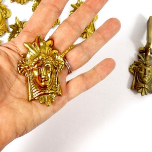 May include: A set of gold-toned metal decorative accents featuring a stylized human face with ornate details. The accents are designed to be used as furniture hardware or decorative elements.