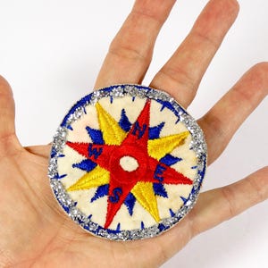 May include: Embroidered compass patch with red, yellow, and blue starburst design. The patch has a white background and a silver glitter border. A decorative nautical item.