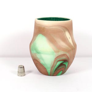 May include: A ceramic vase with a unique marbled design in shades of brown, cream, and green. The interior is a deep green. A small, silver thimble sits beside the vase.