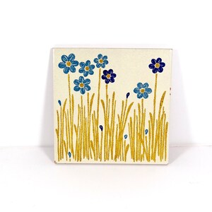 May include: Square ceramic tile with a cream background featuring a floral design. The design includes blue flowers and gold-colored grass. The tile is a decorative piece, suitable for home decor.