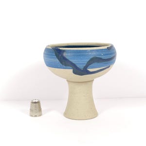 May include: A ceramic chalice with a cream-colored base and a blue-painted rim. The rim features dark blue abstract designs. A small silver thimble is next to the chalice. The chalice is approximately 4 inches tall.