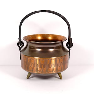 May include: A vintage copper cauldron with a black handle and three small brass feet. The cauldron has a decorative band with an etched design around the middle. The interior of the cauldron is a bright copper color.