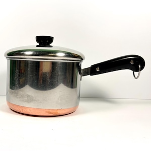 May include: A stainless steel saucepan with a copper-colored base, a matching lid with a black knob, and a black handle featuring a small metal hanging loop. The pot has a polished, reflective finish and a cylindrical shape for cooking tasks.
