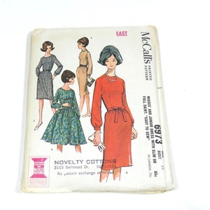 May include: A vintage McCall's sewing pattern for a dress, featuring four models wearing different styles of dresses. The pattern number is 6973 and the pattern is described as "Misses' and Junior Dress with Slim or Full Skirt - Easy to Sew". The pattern is for novelty cottons.