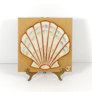 May include: A ceramic tile with a raised seashell design. The shell is white with brown and blue accents. The tile is on a small gold stand. The tile is marked with the letter "J".