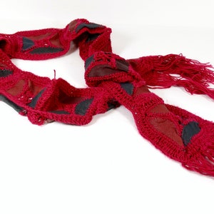 May include: A red crocheted scarf with black leather accents and fringe. The scarf is made of a soft, textured yarn and has a unique, bohemian style.