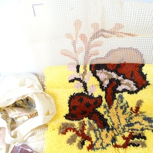 May include: A partially completed rug hooking project with a yellow background and a floral design featuring brown, orange, pink, and gray yarn. The project is on a canvas grid with a bag of yarn and a label that reads "Euclilla Rug Yarn Super Washable"