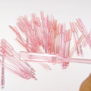 May include: A collection of small, translucent pink plastic applicators, possibly for cosmetic use, are scattered on a white surface. The applicators have a paddle-like end and a thin handle. Some are grouped together, while others are scattered.