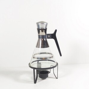 May include: A glass carafe with a silver lid and handle, sitting on a black metal stand with a candle holder. The carafe has a silver band around the middle.