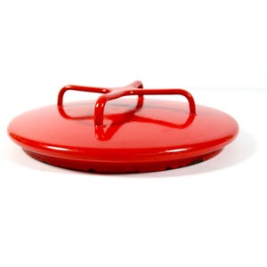 May include: A circular, bright red lid featuring a glossy finish and a central, four-pronged metal handle. The lid has a smooth, slightly domed top surface and a flat, circular rim that extends slightly beyond the main body of the lid.