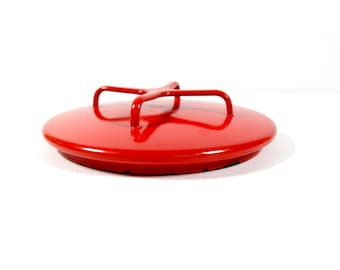 May include: A circular, bright red lid featuring a glossy finish and a central, four-pronged metal handle. The lid has a smooth, slightly domed top surface and a flat, circular rim that extends slightly beyond the main body of the lid.
