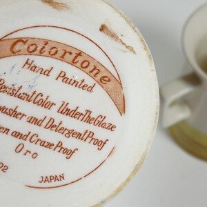 Puede incluir: Una jarra de cer&aacute;mica blanca con un esmalte amarillo y marr&oacute;n. La base de la jarra tiene una etiqueta marr&oacute;n que dice "Colortone Hand Painted Acid Resistant Color Under The Glaze Dishwasher and Detergent Proof Oven and Craze Proof Oro 4702 JAPAN".