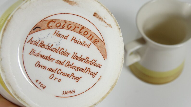 Puede incluir: Una jarra de cer&aacute;mica blanca con un esmalte amarillo y marr&oacute;n. La base de la jarra tiene una etiqueta marr&oacute;n que dice "Colortone Hand Painted Acid Resistant Color Under The Glaze Dishwasher and Detergent Proof Oven and Craze Proof Oro 4702 JAPAN".