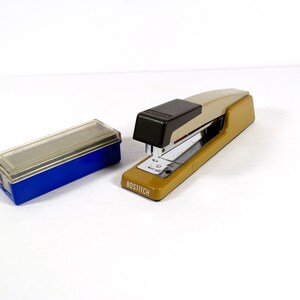 May include: A tan and black Bostitch stapler with a silver base sits next to a blue and clear staple box. The stapler has the word "BOSTITCH" printed on the side. The items are on a white background.