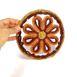 May include: A round, woven trivet with a floral design. The trivet features a central flower with petals in shades of orange, yellow, and brown, surrounded by a dark brown border. The outer edge is a lighter tan color.