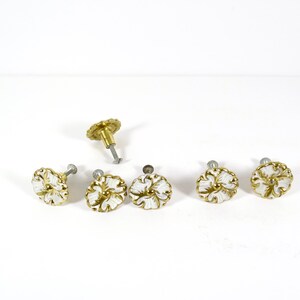 May include: A collection of floral-shaped cabinet knobs. The knobs feature a white and gold color scheme. One knob is disassembled, showing the screw. The knobs are likely for furniture or decorative purposes.