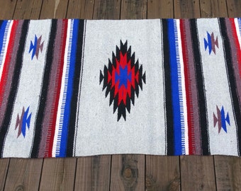May include: A gray woven throw blanket with a geometric pattern in black, red, and blue. The blanket has fringe on the ends.