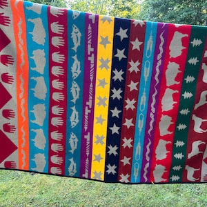 May include: A multi-colored woven blanket featuring vertical stripes with repeating patterns of handprints, fish, stars, feathers, bison, and pine trees. The design includes vibrant bands of red, orange, blue, yellow, dark blue, and green with a black border.