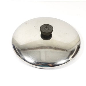 May include: A stainless steel pot lid with a dark, round handle. The lid is circular and reflective, with a slightly domed shape. The handle appears to be made of a dark, possibly heat-resistant material. The lid is designed for cookware.