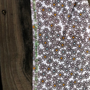 Puede incluir: Primer plano de una tela con fondo blanco cubierta de peque&ntilde;as flores de margarita grises con centros naranjas y celeste. El borde de la tela tiene un borde verde y blanco con texto.